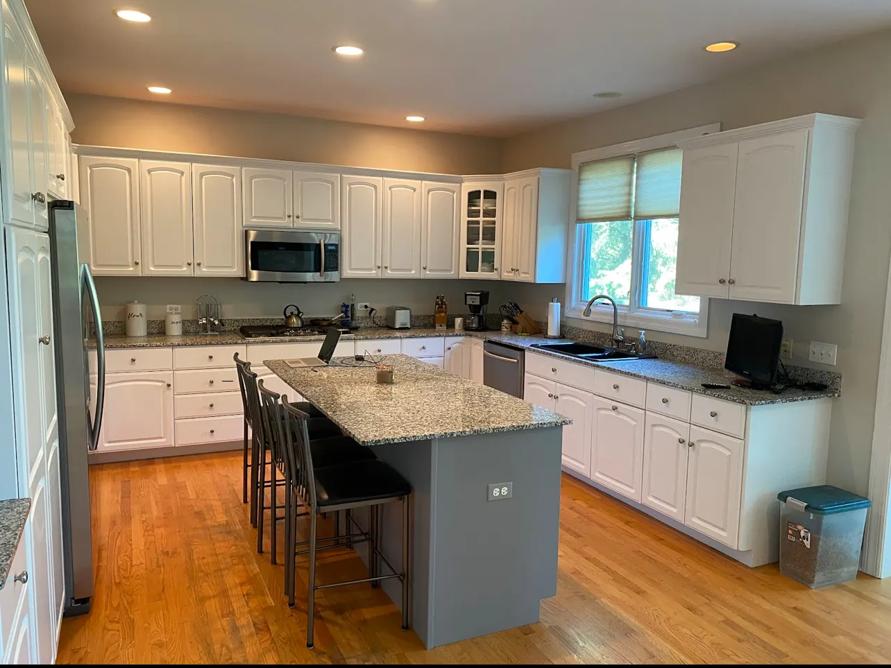 A kitchen with white painted raised-panel cabinets and crown molding, speckled granite countertops, a gray painted island with bar stools, stainless steel appliances and hardwood floors.