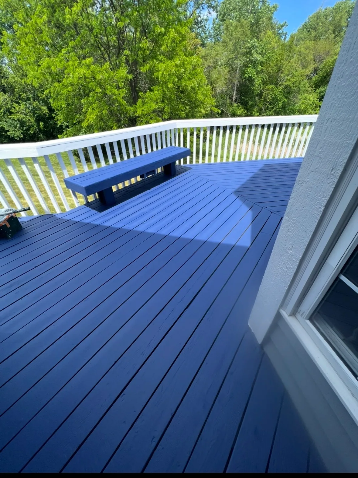 A backyard deck with boards and a bench painted deep navy blue, white painted railings and balusters, and green trees beyond.