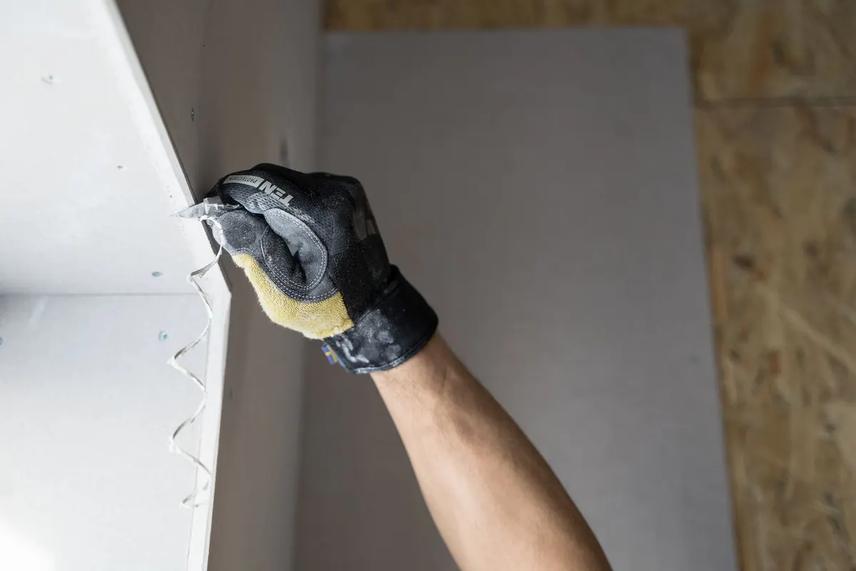 A gloved hand working along the top edge of a drywall sheet where it meets the ceiling, smoothing back a ridge of dried compound.