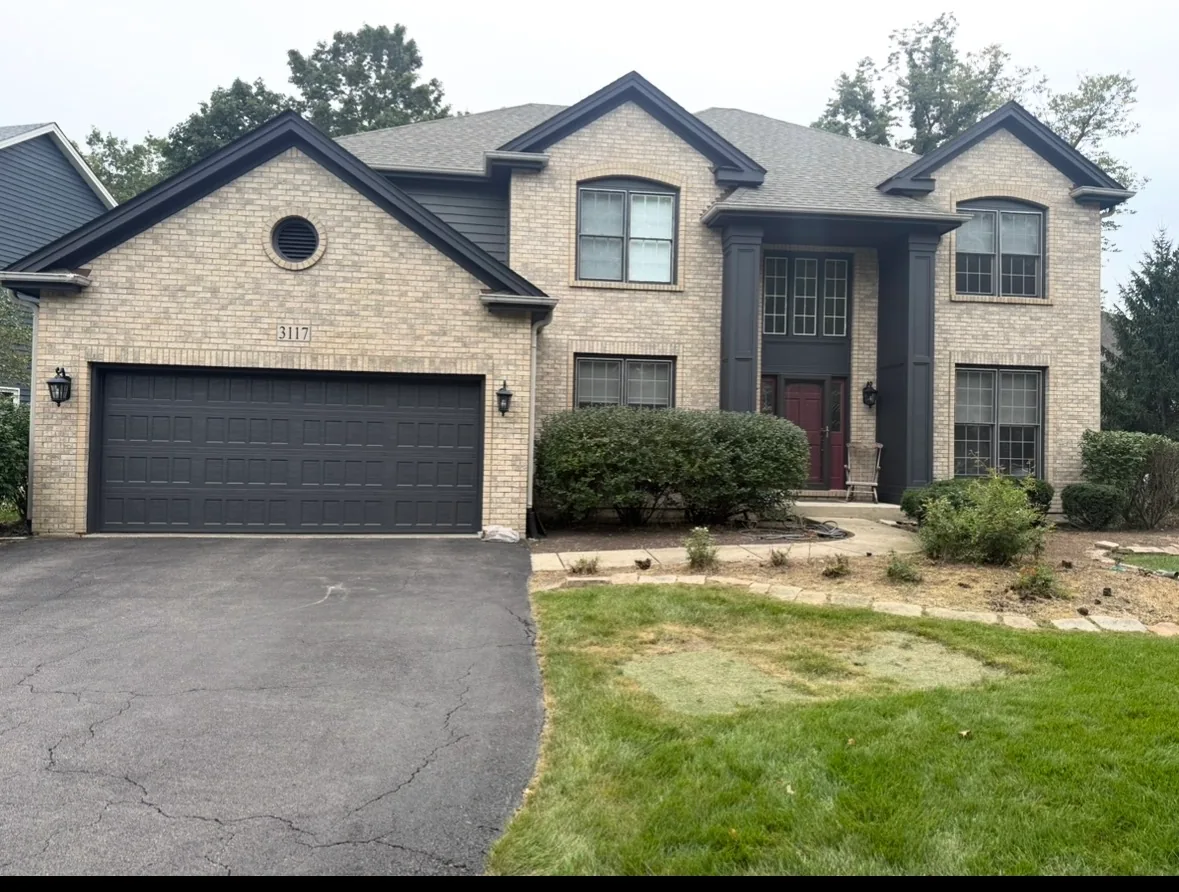 The front of a two-story light tan brick home with charcoal gray painted gables, window trim, entry columns and garage door, a deep red front door, and an asphalt driveway.