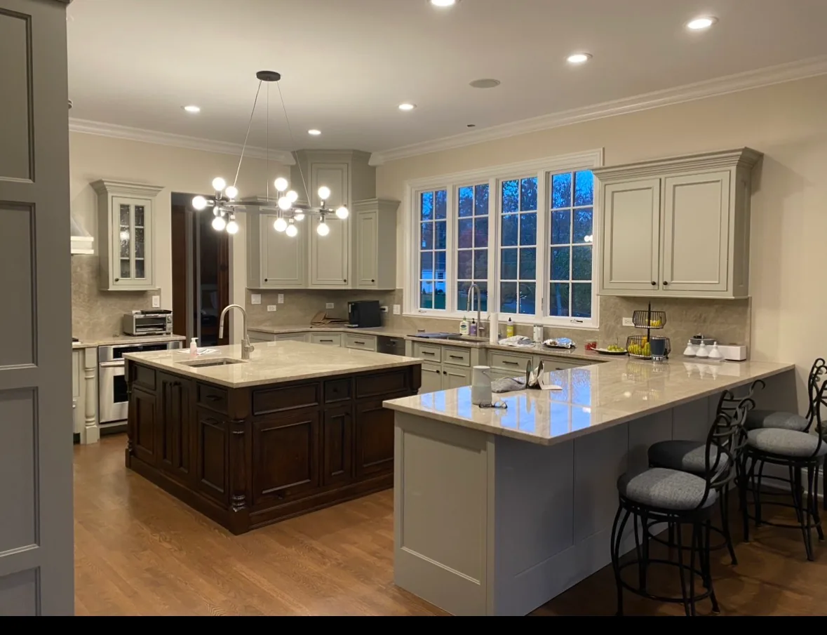 A large kitchen with soft sage-gray painted cabinets, white crown molding and window trim, a dark stained wood island with a pale stone countertop and sink, and a seating peninsula with bar stools over hardwood floors.