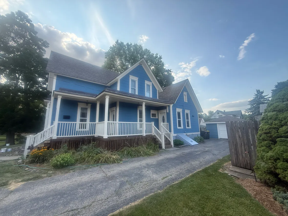 A two-story house with blue painted siding and white trim, a covered front porch with white columns and railings, and a paved driveway running alongside the house to a detached garage.