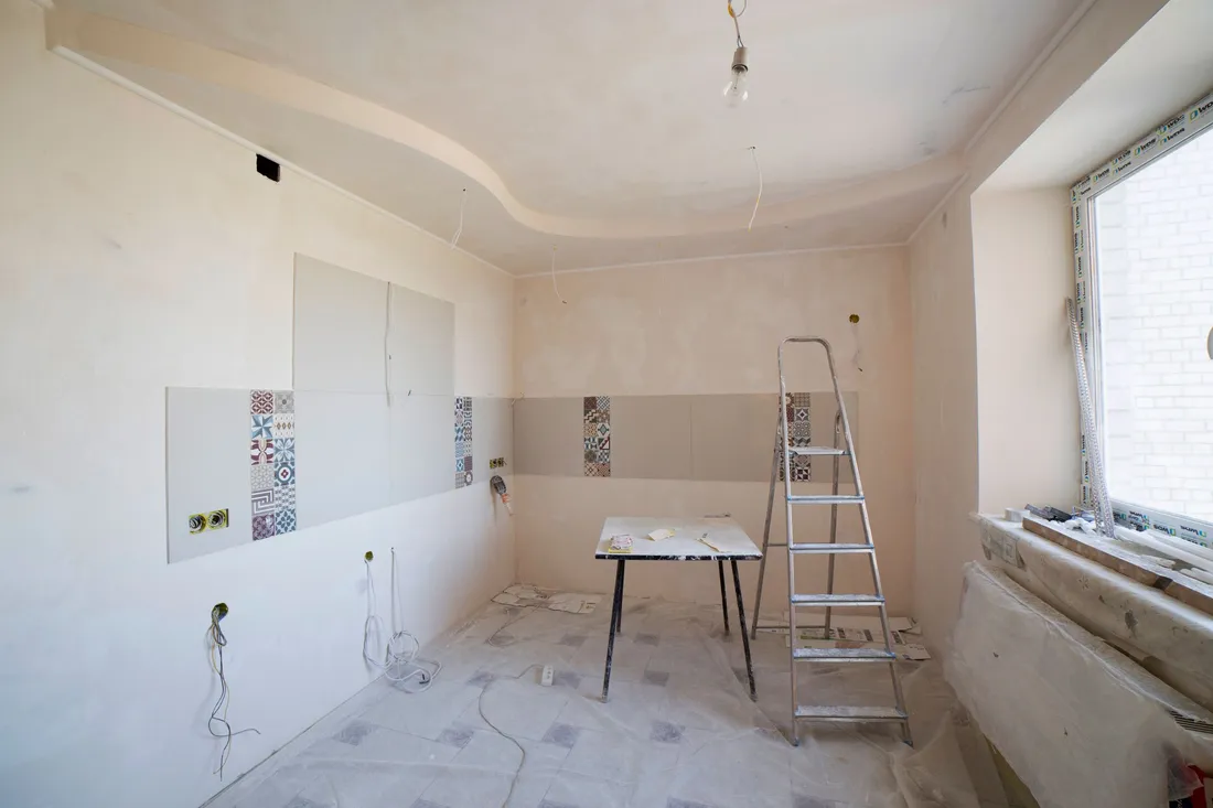 A room part-way through a remodel: prepped bare walls, a step ladder, and drop sheets covering the floor.